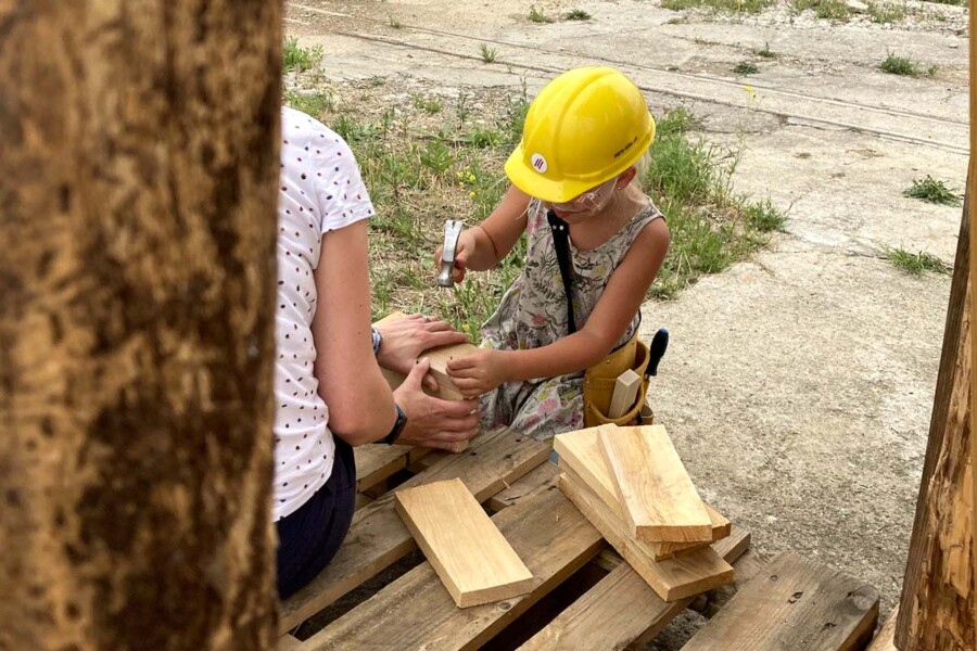 Mädchen schlägt Nagel in ein Holzstück, das von einer Frau mit zwei Händen fest gehalten wird